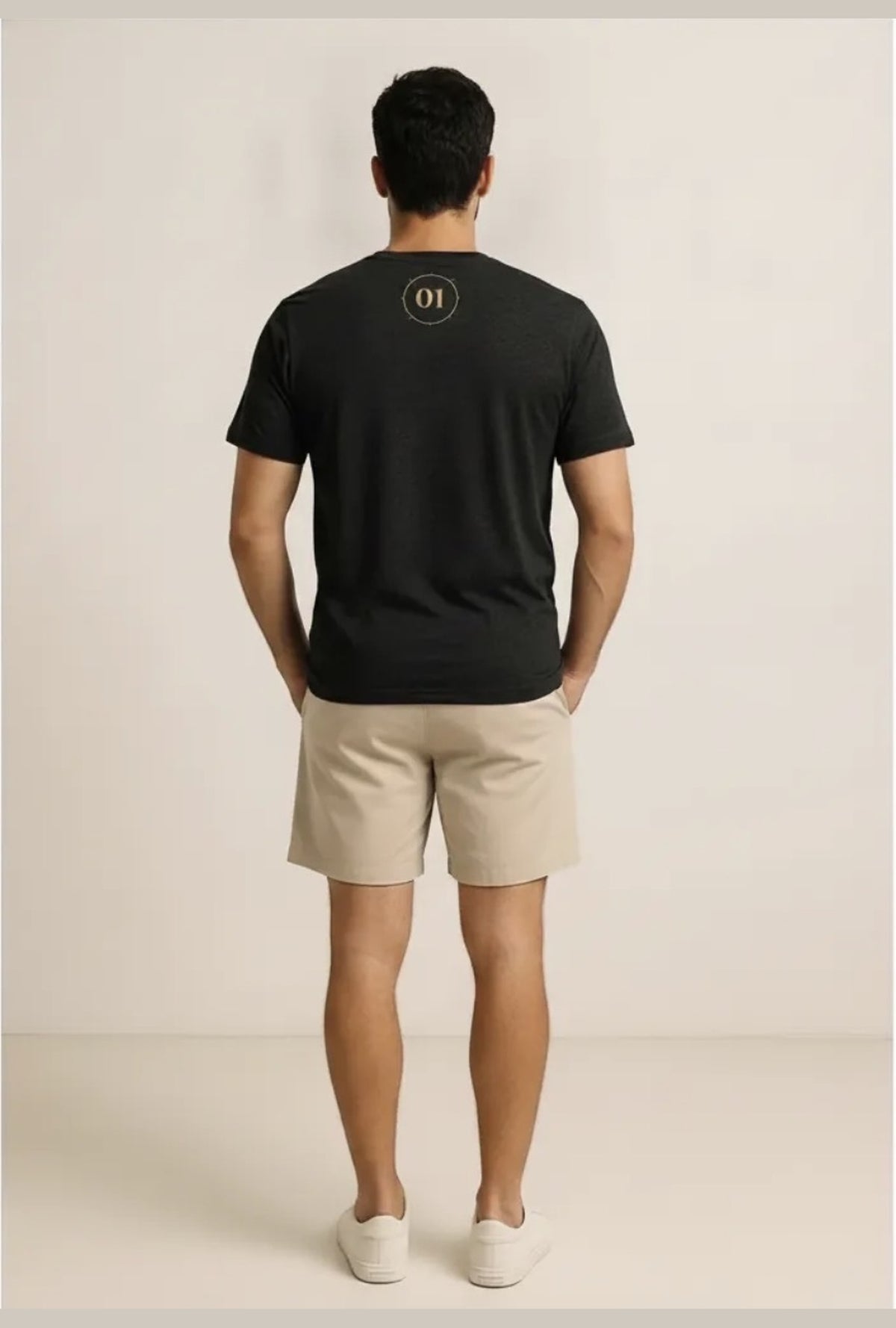 A man viewed from behind wearing a black short-sleeve t-shirt with a small circular logo on the upper back, paired with beige shorts and white sneakers, standing against a plain light beige studio background.